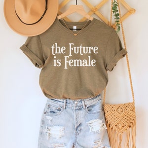 May include: A green t-shirt with the text "the Future is Female" in white lettering. The t-shirt is hanging on a wooden hanger with a straw bag and a brown hat.