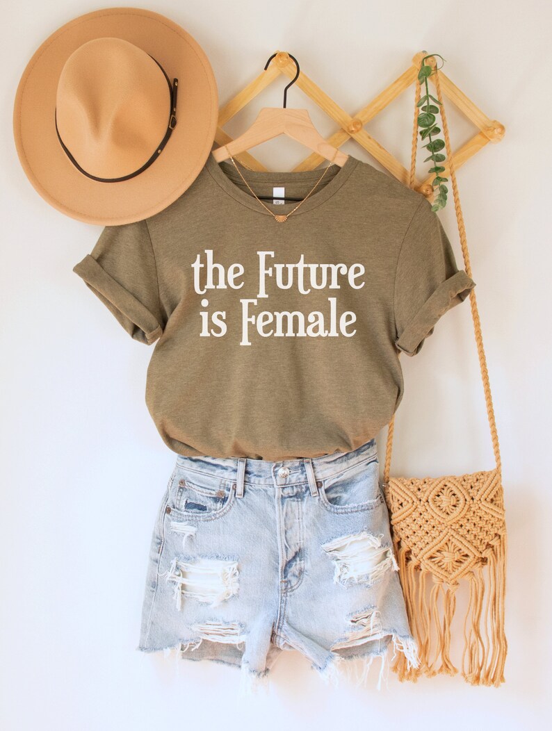May include: A green t-shirt with the text "the Future is Female" in white lettering. The t-shirt is hanging on a wooden hanger with a straw bag and a brown hat.