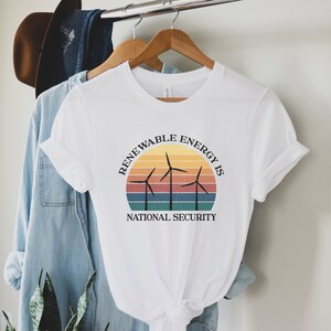 May include: White t-shirt with a graphic of a sunset and wind turbines. The text reads "Renewable Energy is National Security".