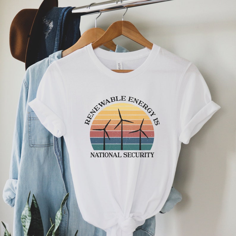 May include: White t-shirt with a graphic of a sunset and wind turbines. The text reads "Renewable Energy is National Security".