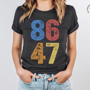 May include: A dark grey t-shirt with the numbers 86 and 47 printed in a vintage style. The numbers are in different colors: 8 is blue, 6 is red, 4 is orange, and 7 is yellow.
