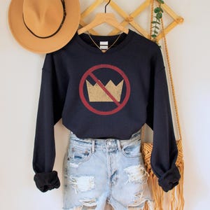 A navy blue unisex crew neck long sleeve fleece-lined sweatshirt with an image of a yellow crown inside a red circle with a slash over it, meaning "no kings". The image has a vintage/distressed look.
