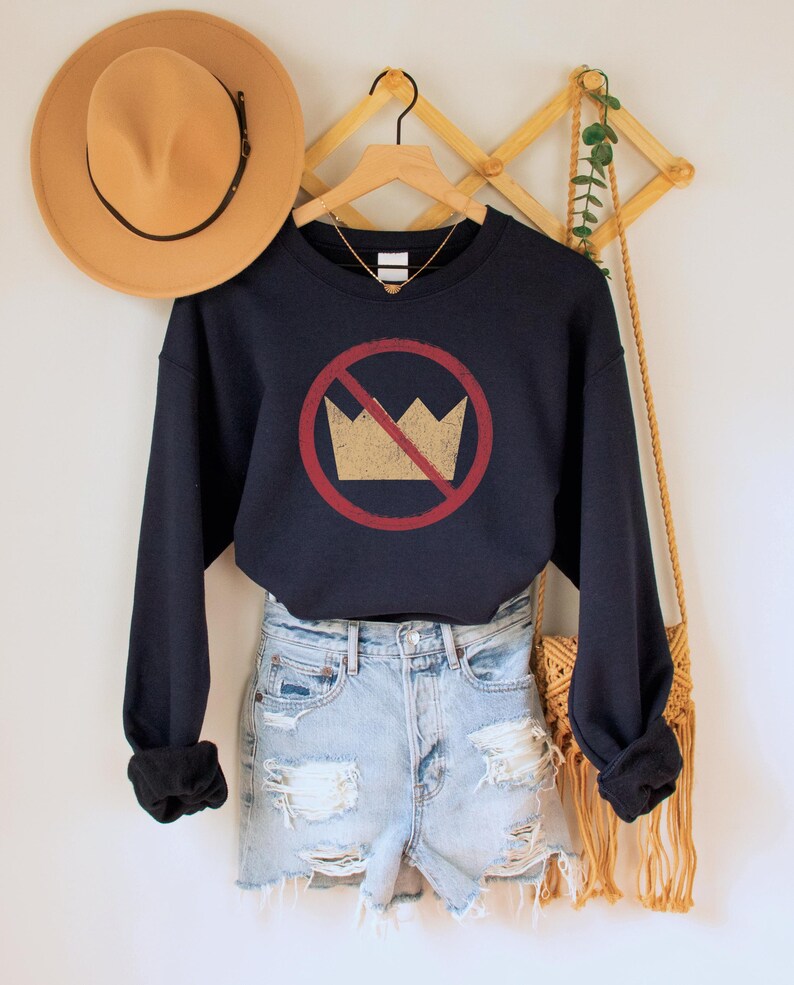 A navy blue unisex crew neck long sleeve fleece-lined sweatshirt with an image of a yellow crown inside a red circle with a slash over it, meaning "no kings". The image has a vintage/distressed look.