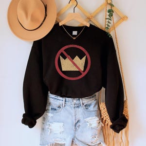 A black unisex crew neck long sleeve fleece-lined sweatshirt with an image of a yellow crown inside a red circle with a slash over it, meaning "no kings". The image has a vintage/distressed look.