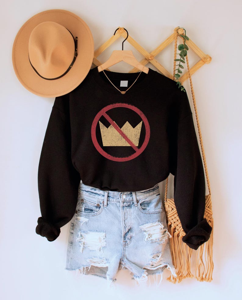 A black unisex crew neck long sleeve fleece-lined sweatshirt with an image of a yellow crown inside a red circle with a slash over it, meaning "no kings". The image has a vintage/distressed look.