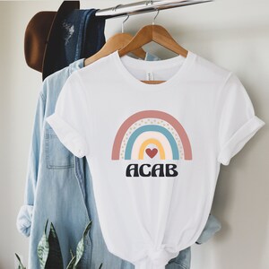 May include: White t-shirt with a rainbow graphic and the text "ACAB" in black letters. The rainbow is made up of pink, blue, yellow, and white stripes with gold stars.