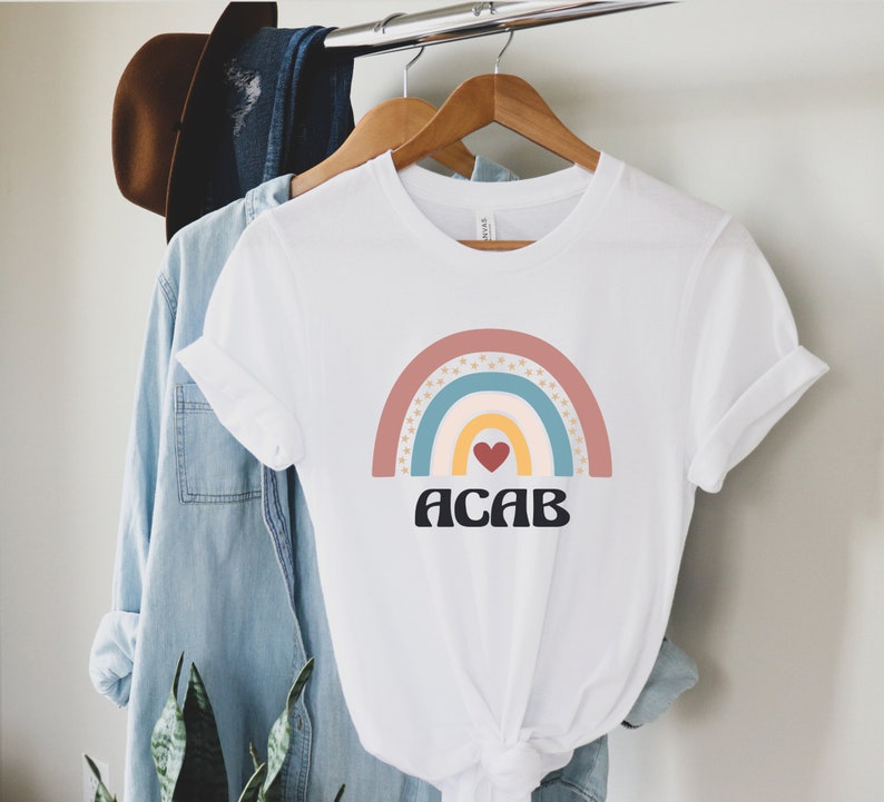 May include: White t-shirt with a rainbow graphic and the text "ACAB" in black letters. The rainbow is made up of pink, blue, yellow, and white stripes with gold stars.