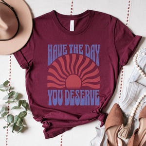 A maroon unisex short sleeve crew neck t-shirt that says "have the day you deserve" on the front in distressed blue retro capitalized font around a distressed image of a red wavy retro sunset