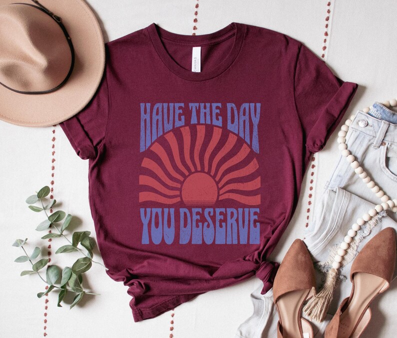 A maroon unisex short sleeve crew neck t-shirt that says "have the day you deserve" on the front in distressed blue retro capitalized font around a distressed image of a red wavy retro sunset