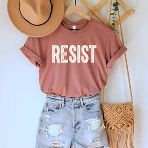 A heather mauve unisex crew neck short sleeve Bella+Canvas t-shirt that says "RESIST" in distressed white tall block text