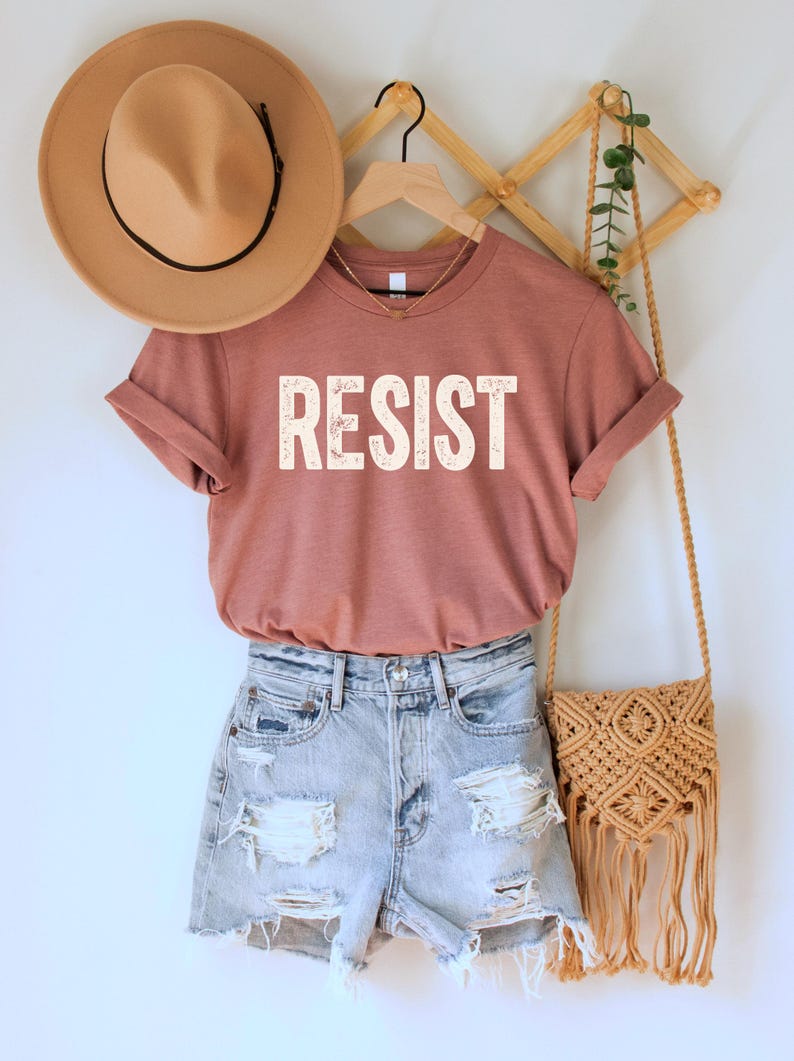 A heather mauve unisex crew neck short sleeve Bella+Canvas t-shirt that says "RESIST" in distressed white tall block text