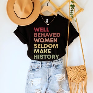 May include: Black t-shirt with the text "WELL BEHAVED WOMEN SELDOM MAKE HISTORY" in a gradient of pink, orange, and yellow. The shirt is on a hanger with a brown hat and a straw bag.