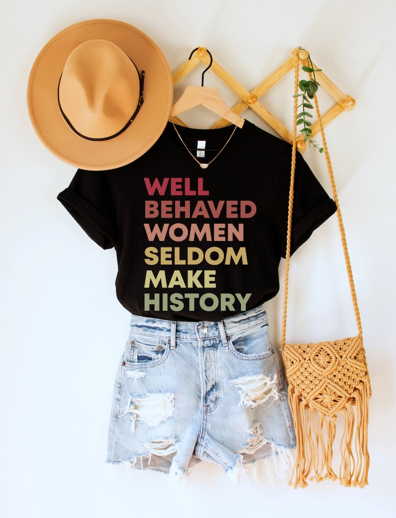 May include: Black t-shirt with the text "WELL BEHAVED WOMEN SELDOM MAKE HISTORY" in a gradient of pink, orange, and yellow. The shirt is on a hanger with a brown hat and a straw bag.
