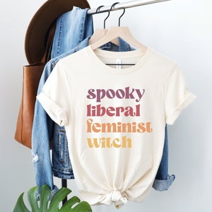A natural (light beige) unisex crew neck t-shirt with the phrase "spooky liberal feminist witch" in purple, red, orange, and yellow text