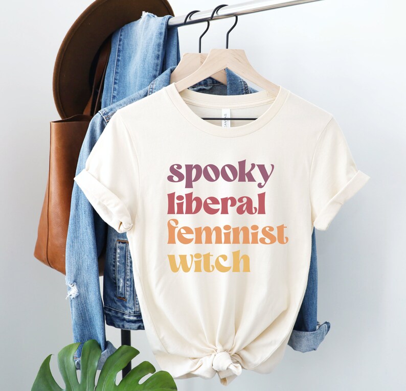 A natural (light beige) unisex crew neck t-shirt with the phrase "spooky liberal feminist witch" in purple, red, orange, and yellow text