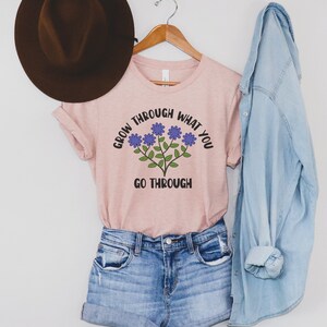 May include: A light pink t-shirt with the text "GROW THROUGH WHAT YOU GO THROUGH" and a floral design. The shirt is paired with denim shorts, a denim shirt, and a brown hat.