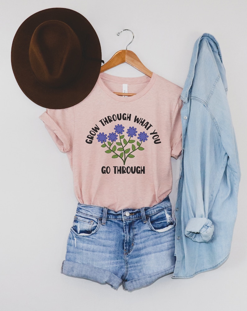 May include: A light pink t-shirt with the text "GROW THROUGH WHAT YOU GO THROUGH" and a floral design. The shirt is paired with denim shorts, a denim shirt, and a brown hat.