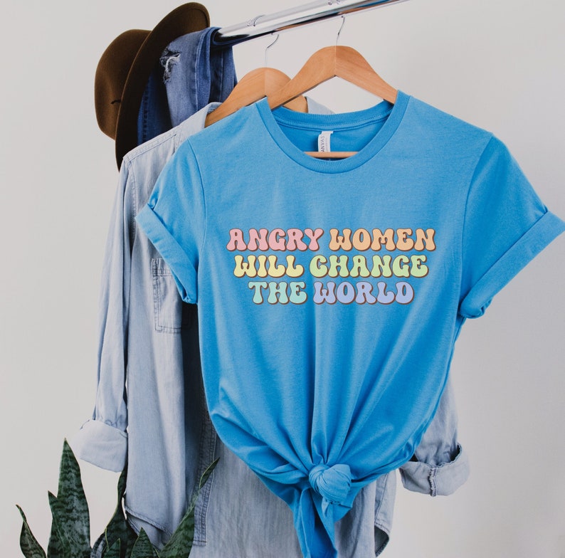 Może przedstawiać: Jasnoniebieska koszulka z napisem "Angry Women Will Change The World" w kolorach tęczy.