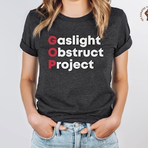 May include: Dark gray t-shirt with the words "Gaslight, GOP, obstruct, project" in white and red lettering. The shirt has rolled-up sleeves and is paired with blue jeans. A small logo is visible in the upper right corner.