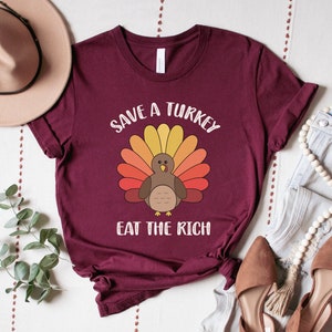 K&ouml;nnte beinhalten: Burgunderfarbenes T-Shirt mit einer Cartoon-Truthahn-Illustration mit bunten Federn. Der Text "SAVE A TURKEY" ist &uuml;ber dem Truthahn gew&ouml;lbt, darunter steht "EAT THE RICH". Das Shirt wird mit Jeans, Schuhen und einem Hut pr&auml;sentiert.