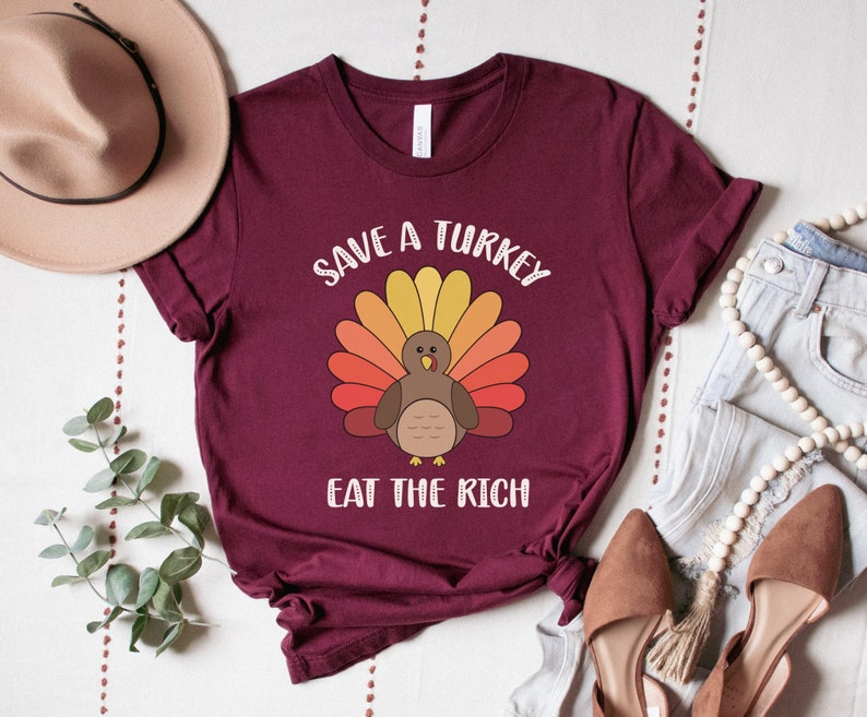 K&ouml;nnte beinhalten: Burgunderfarbenes T-Shirt mit einer Cartoon-Truthahn-Illustration mit bunten Federn. Der Text "SAVE A TURKEY" ist &uuml;ber dem Truthahn gew&ouml;lbt, darunter steht "EAT THE RICH". Das Shirt wird mit Jeans, Schuhen und einem Hut pr&auml;sentiert.