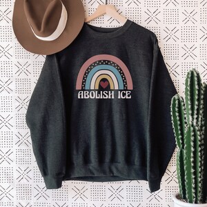 May include: A dark grey sweatshirt with a rainbow graphic and the text "ABOLISH ICE" in white. The rainbow has a red heart in the center.