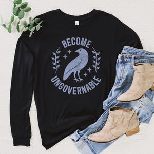 A black unisex long sleeve crew neck t-shirt that says  "BECOME UNGOVERNABLE" on the chest in block text around an image of a crow, leaves, and retro stars. The whole graphic and text are light blue and have a distressed/vintage look.