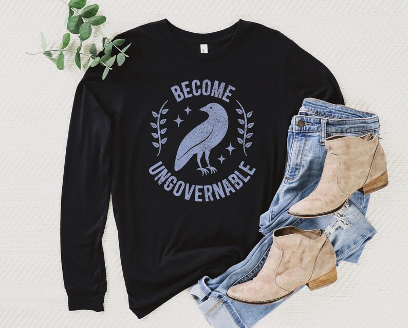A black unisex long sleeve crew neck t-shirt that says  "BECOME UNGOVERNABLE" on the chest in block text around an image of a crow, leaves, and retro stars. The whole graphic and text are light blue and have a distressed/vintage look.