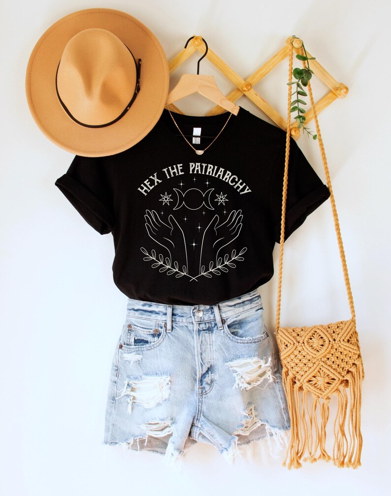 A black unisex crew neck short sleeve Bella+Canvas t-shirt that says "hex the patriarchy" in a white uppercase distressed font, above a witchy white graphic with leaves, hands, stars, and a three moon symbol