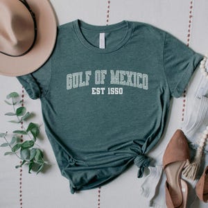 A heather forest green unisex crew neck short sleeve Bella+Canvas brand t-shirt that says "GULF OF MEXICO" on the chest in white outline college style font with white stripes inside the letters. The text is curved over smaller solid white letters in the same font that say "EST 1550". The graphic has a distressed/vintage look