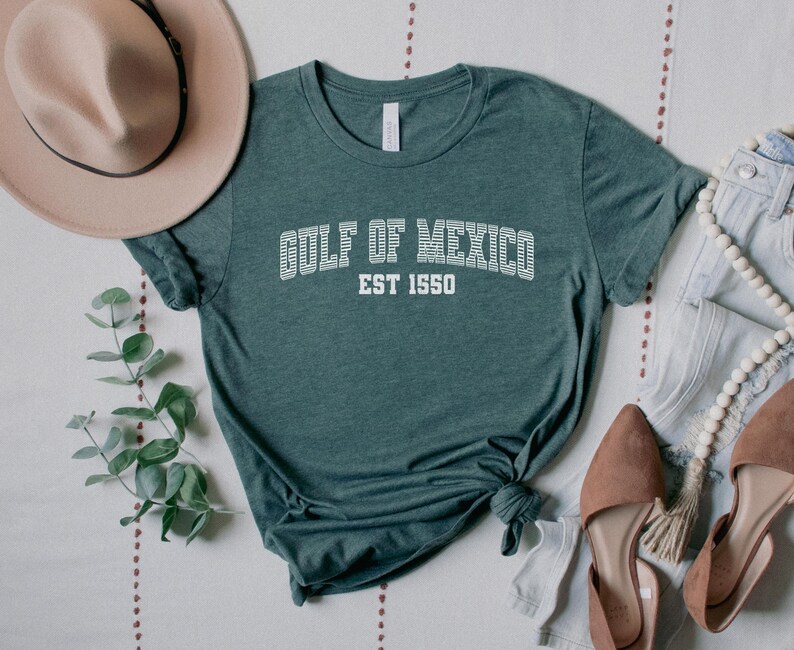 A heather forest green unisex crew neck short sleeve Bella+Canvas brand t-shirt that says "GULF OF MEXICO" on the chest in white outline college style font with white stripes inside the letters. The text is curved over smaller solid white letters in the same font that say "EST 1550". The graphic has a distressed/vintage look