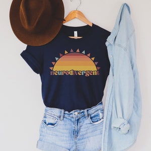 May include: A navy blue t-shirt with a sunset graphic and the text "neurodivergent". The shirt is hanging on a wooden hanger with a brown hat and a light blue denim shirt on either side.