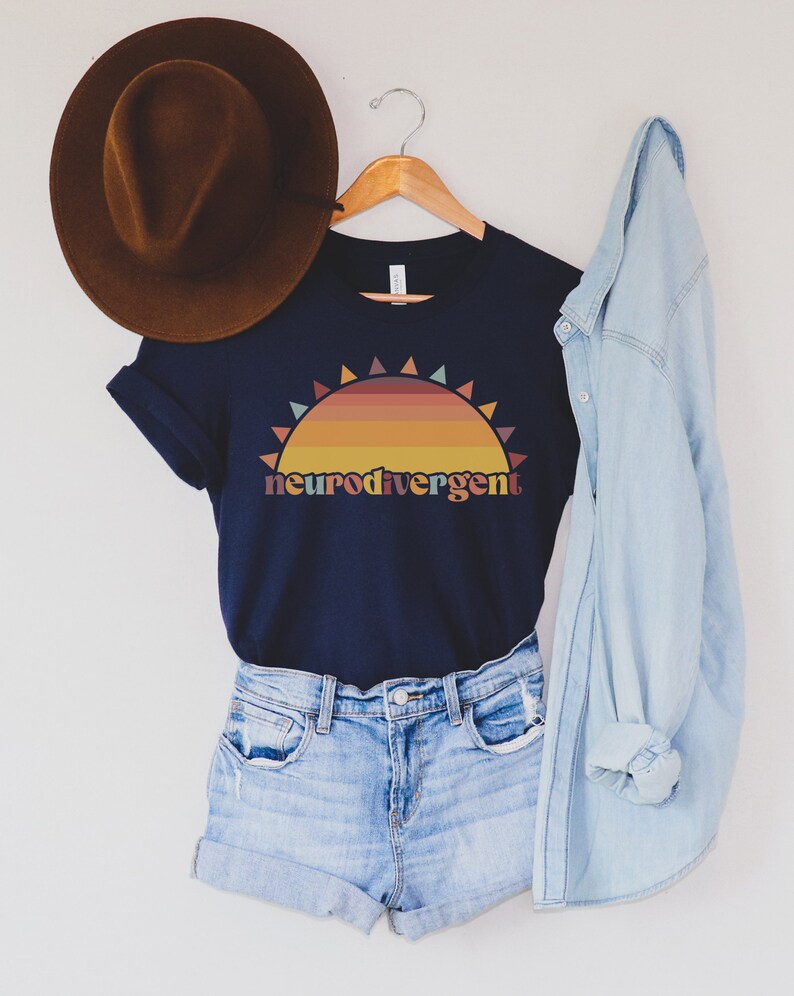 May include: A navy blue t-shirt with a sunset graphic and the text "neurodivergent". The shirt is hanging on a wooden hanger with a brown hat and a light blue denim shirt on either side.