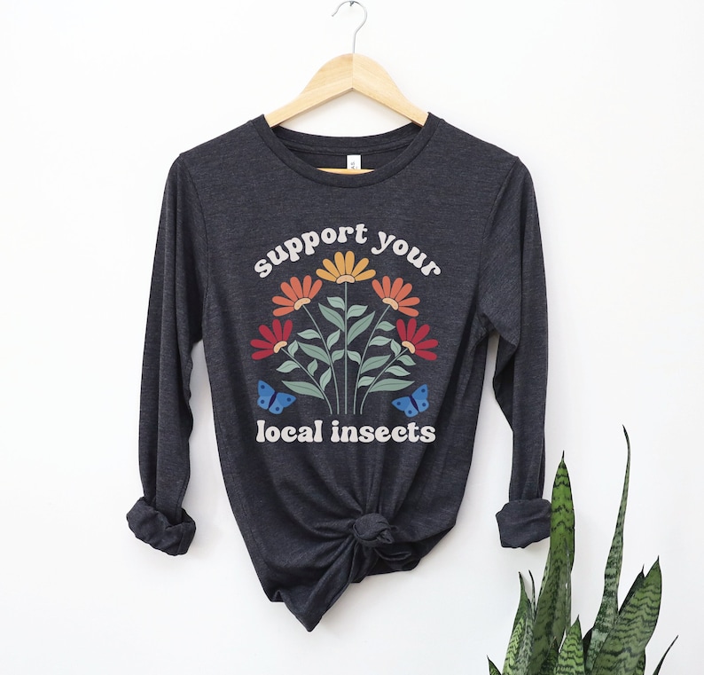 A dark grey heather unisex long sleeve crew neck t-shirt with the words "support your local insects" in white text around an image of colorful flowers and butterflies