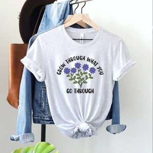 May include: A light gray t-shirt with the text "GROW THROUGH WHAT YOU GO THROUGH" in black and a floral design with purple flowers and green stems. The shirt is hanging on a wooden hanger.