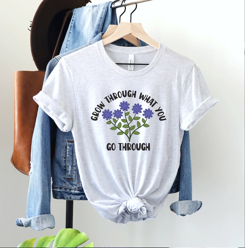 May include: A light gray t-shirt with the text "GROW THROUGH WHAT YOU GO THROUGH" in black and a floral design with purple flowers and green stems. The shirt is hanging on a wooden hanger.