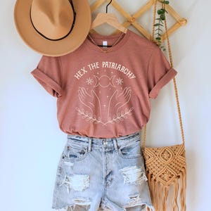 A heather mauve unisex crew neck short sleeve Bella+Canvas t-shirt that says "hex the patriarchy" in a white uppercase distressed font, above a witchy white graphic with leaves, hands, stars, and a three moon symbol