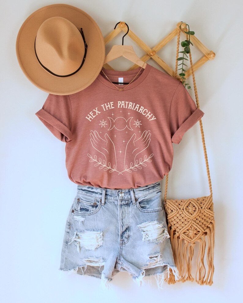 A heather mauve unisex crew neck short sleeve Bella+Canvas t-shirt that says "hex the patriarchy" in a white uppercase distressed font, above a witchy white graphic with leaves, hands, stars, and a three moon symbol