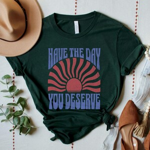 A forest green unisex short sleeve crew neck t-shirt that says "have the day you deserve" on the front in distressed blue retro capitalized font around a distressed image of a red wavy retro sunset