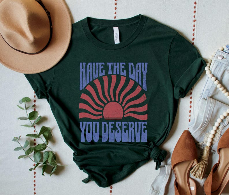A forest green unisex short sleeve crew neck t-shirt that says "have the day you deserve" on the front in distressed blue retro capitalized font around a distressed image of a red wavy retro sunset