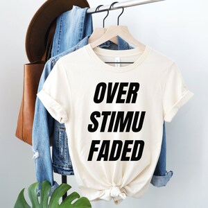 May include: Cream-colored t-shirt with the words "OVER STIMU FADED" in bold black letters. The shirt is hanging on a wooden hanger, alongside a denim jacket, a brown leather tote bag, and a brown hat.