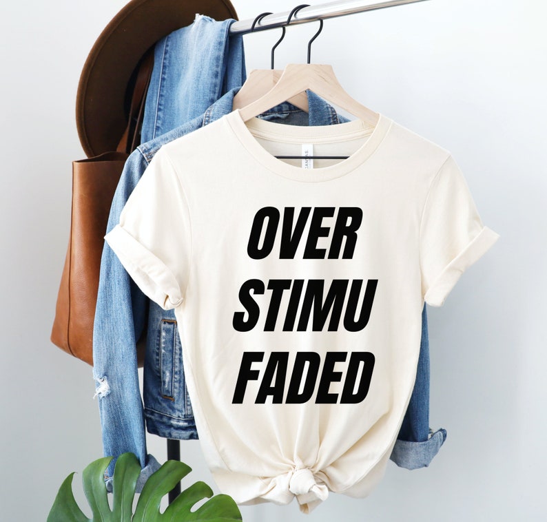 May include: Cream-colored t-shirt with the words "OVER STIMU FADED" in bold black letters. The shirt is hanging on a wooden hanger, alongside a denim jacket, a brown leather tote bag, and a brown hat.