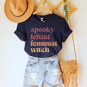 May include: A navy blue t-shirt with the text "spooky leftist feminist witch" in a gradient of pink, orange, and yellow. The shirt is hanging on a wooden rack with a straw hat and a woven bag.