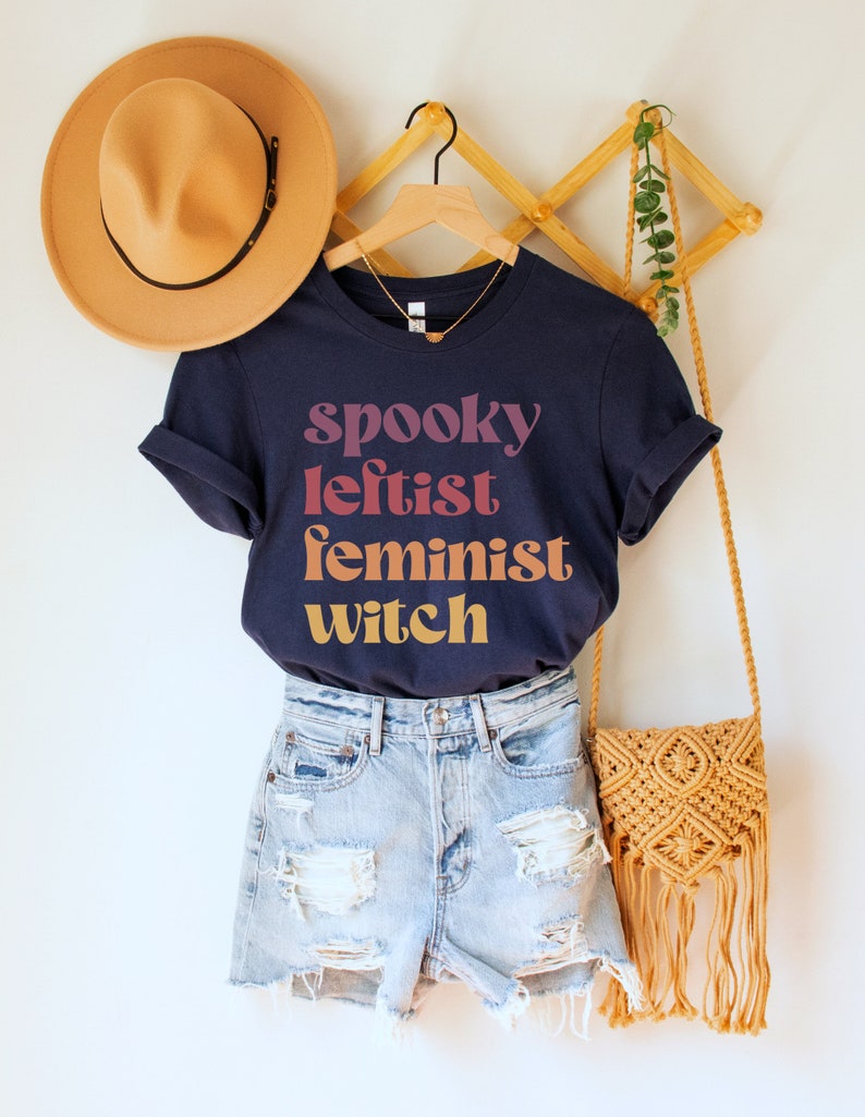 May include: A navy blue t-shirt with the text "spooky leftist feminist witch" in a gradient of pink, orange, and yellow. The shirt is hanging on a wooden rack with a straw hat and a woven bag.