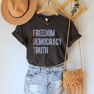 A dark grey heather unisex crew neck short sleeve Bella+Canvas t-shirt that says "FREEDOM DEMOCRACY TRUTH" on the chest in three lines in blue block text with white stars inside the letters. The first letter of each word in in red with white stars, subtly spelling "FDT" down the shirt
