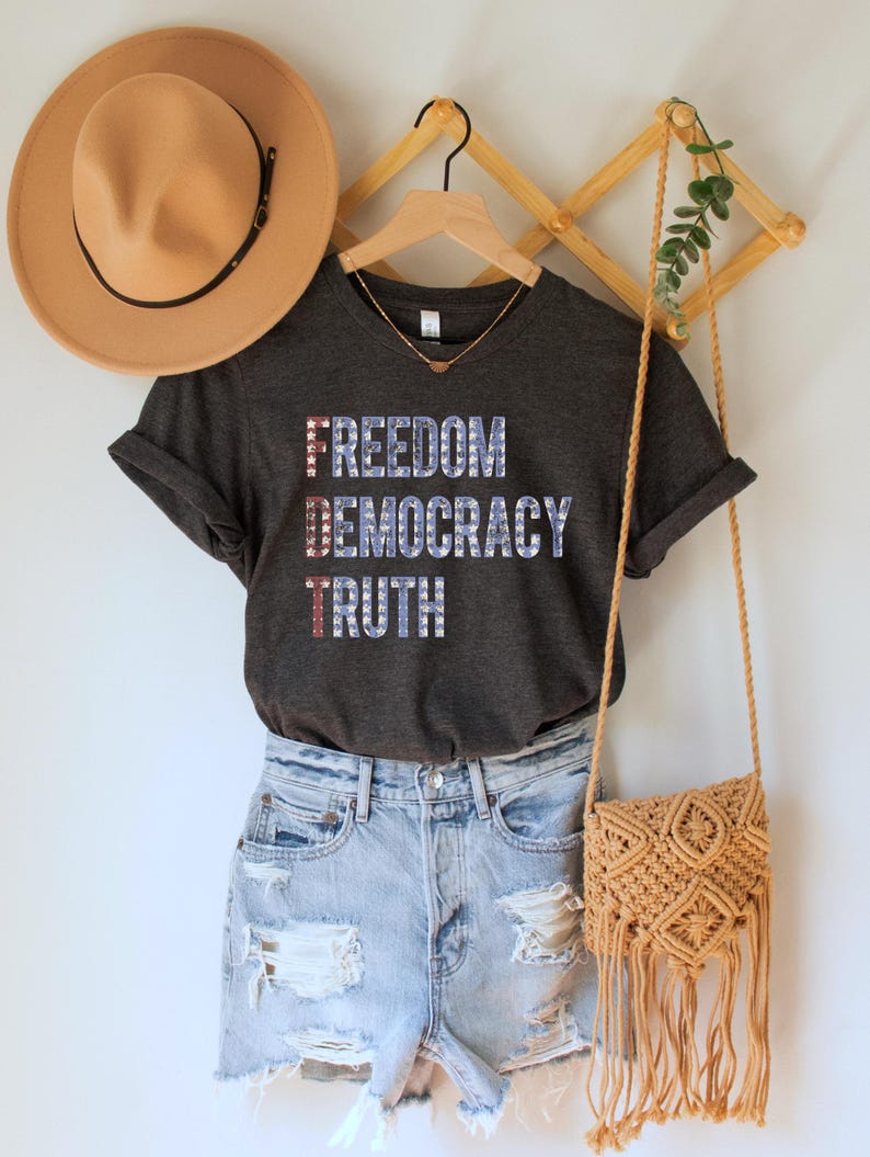A dark grey heather unisex crew neck short sleeve Bella+Canvas t-shirt that says "FREEDOM DEMOCRACY TRUTH" on the chest in three lines in blue block text with white stars inside the letters. The first letter of each word in in red with white stars, subtly spelling "FDT" down the shirt