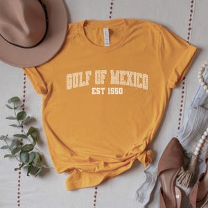 A gold (dark yellow) unisex crew neck short sleeve Bella+Canvas brand t-shirt that says "GULF OF MEXICO" on the chest in white outline college style font with white stripes inside the letters. The text is curved over smaller solid white letters in the same font that say "EST 1550". The graphic has a distressed/vintage look
