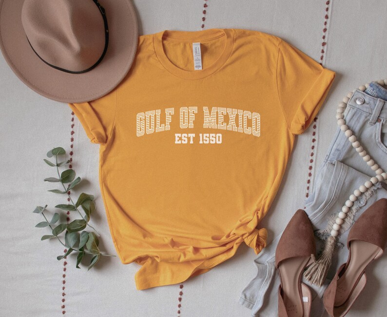 A gold (dark yellow) unisex crew neck short sleeve Bella+Canvas brand t-shirt that says "GULF OF MEXICO" on the chest in white outline college style font with white stripes inside the letters. The text is curved over smaller solid white letters in the same font that say "EST 1550". The graphic has a distressed/vintage look