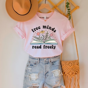 A pink unisex crew neck short sleeve t-shirt with black text saying "free minds read freely" around a graphic of a book with colorful flowers and stars coming out of it