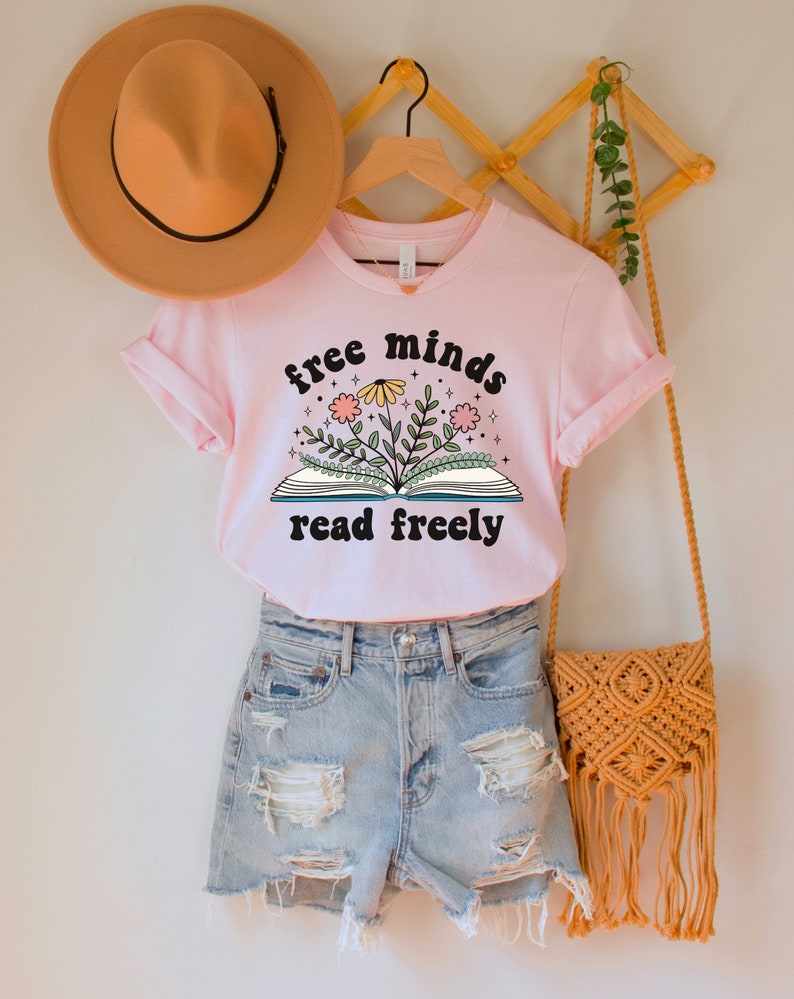 A pink unisex crew neck short sleeve t-shirt with black text saying "free minds read freely" around a graphic of a book with colorful flowers and stars coming out of it